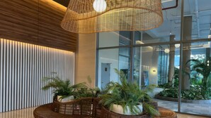 A modern lobby with a large glass window, a woven ceiling light fixture, and a seating area with wicker furniture and potted plants. - Diamond Island - Mekong Retreat Apartment (Ho Chi Minh City)