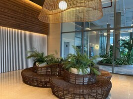 Un vestíbulo moderno con un amplio ventanal, una lámpara de techo trenzada y una zona de estar con muebles de mimbre y plantas en macetas.