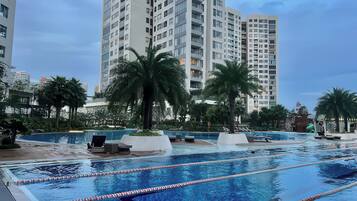 A swimming pool with lane dividers, surrounded by palm trees and lounge chairs, with tall residential buildings in the background.