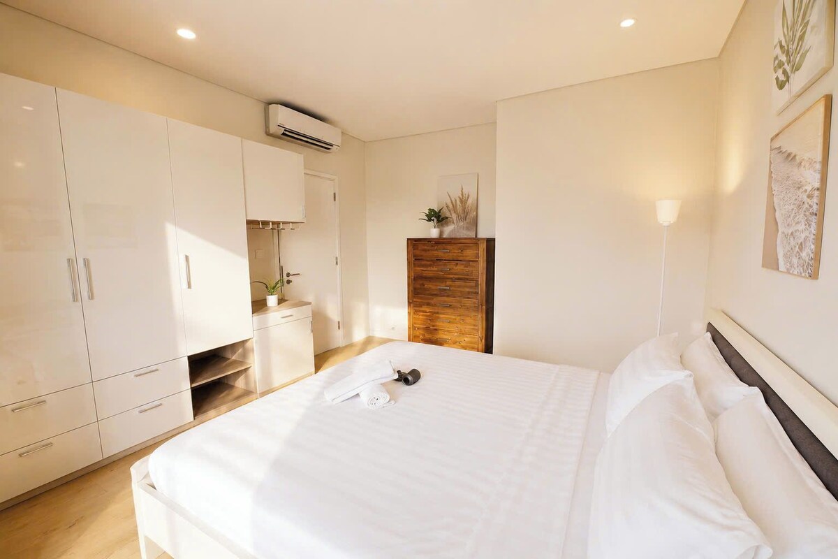 A bedroom with a bed, white curtains, a wooden dresser, and a wall-mounted air conditioner.
