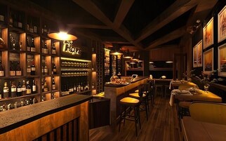 Una zona de bar con estantes de madera llenos de botellas, un mostrador con taburetes y cuadros en las paredes.