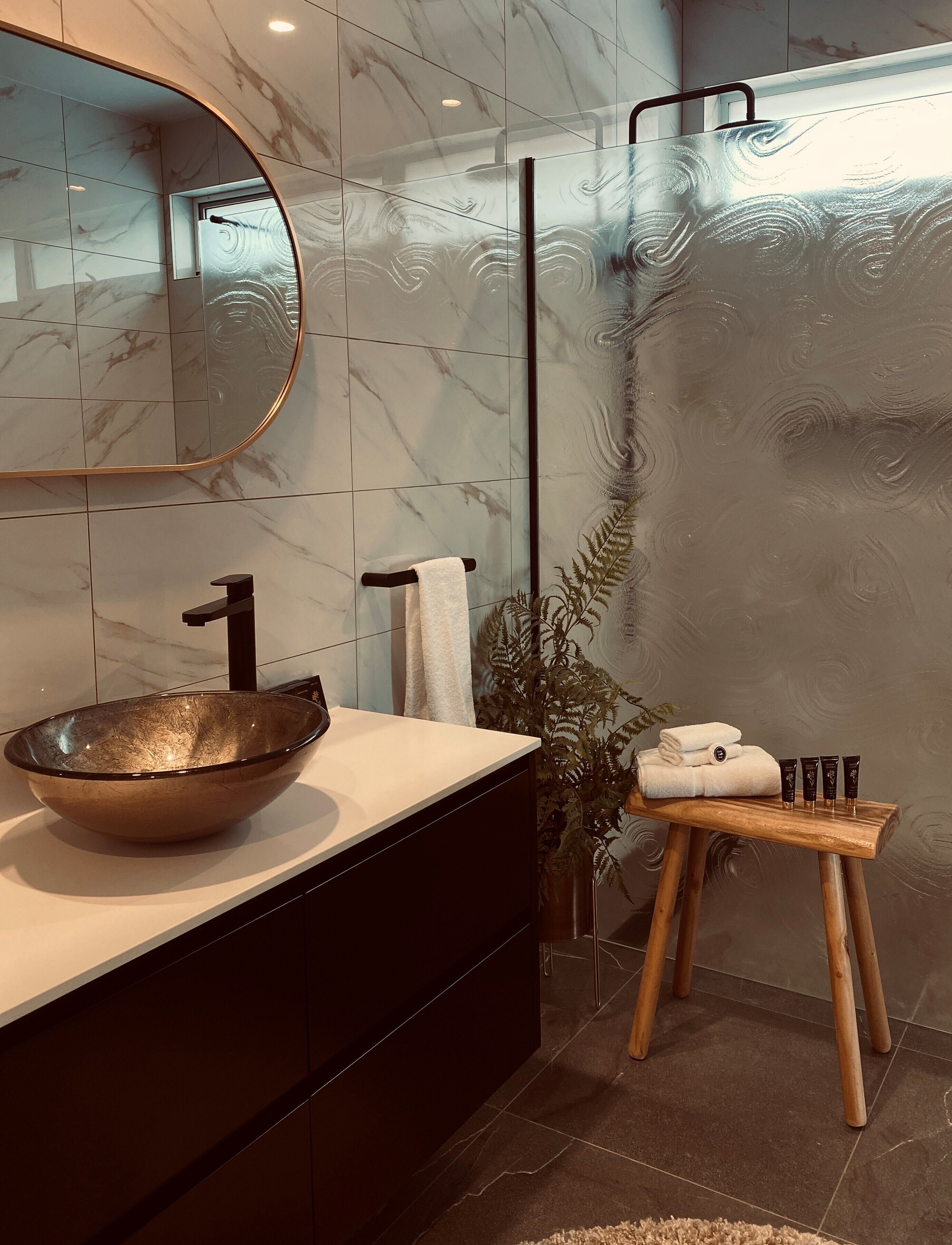 A modern bathroom with a large mirror, a glass bowl sink, and a wooden stool with toiletries.