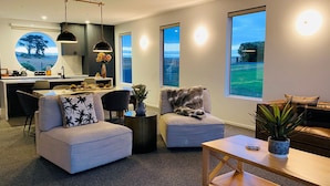 A modern living room with a sofa, armchair, and coffee table. There is a dining area with a table and chairs, and a kitchen in the background. Large windows provide views of the outdoors. - Rose's Villa (Stanley)
