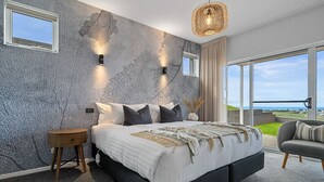 A modern bedroom with a large bed, a nightstand, a chair, and a view of the sea through large windows. - Rose's Villa (Stanley)