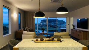 A modern hotel room with a large window offering a scenic view of a mountain and town. There is a dining area with a marble table, a sofa, and a TV mounted on the wall. - Rose's Villa (Stanley)