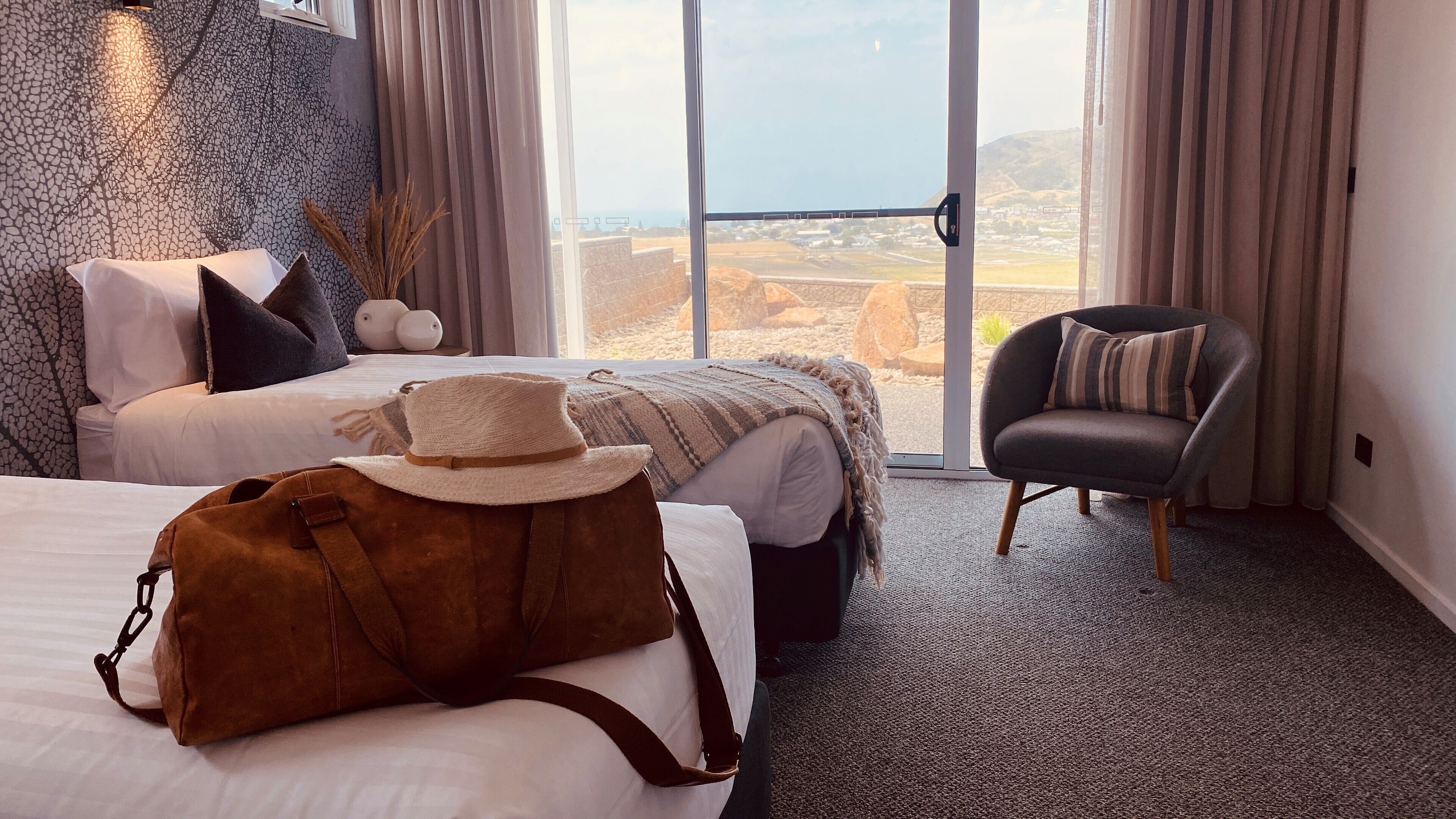A hotel room with two beds, a large window with a view of the countryside, a chair, and a brown bag on one bed.