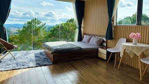 A bedroom with a large bed, a small table with a vase of flowers, and a view of the landscape. - Windy Hill Homestay (Xuan Truong - Da Lat)