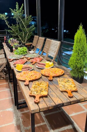 A wooden table with various dishes and drinks, surrounded by chairs and potted plants. - Windy Hill Homestay (Xuan Truong - Da Lat)