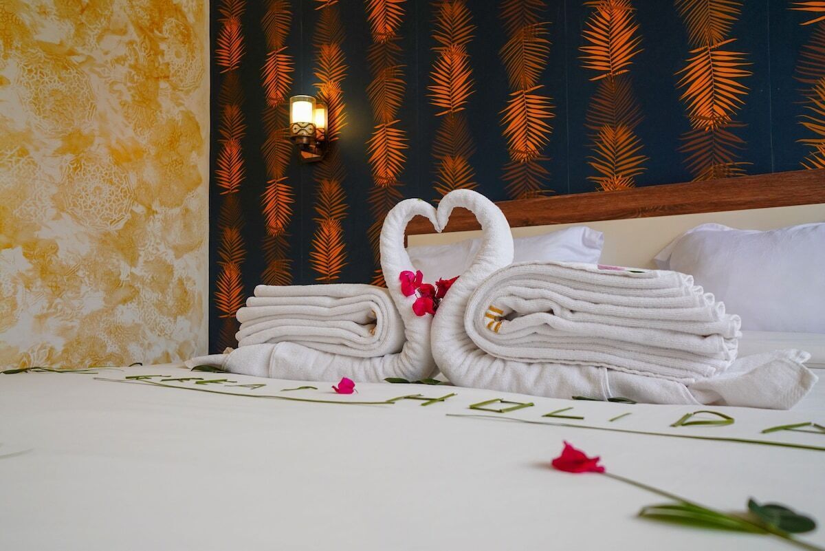 A neatly made bed with folded towels and a heart-shaped towel arrangement, a wall with a palm leaf pattern, and a wall-mounted light fixture.