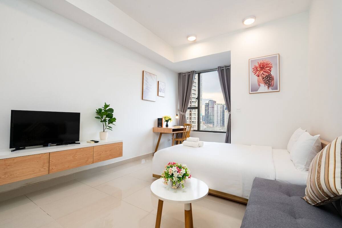 Una habitación de hotel moderna con una cama grande, un televisor sobre un mueble de madera, una mesita con un florero y vistas a la ciudad a través de la ventana.
