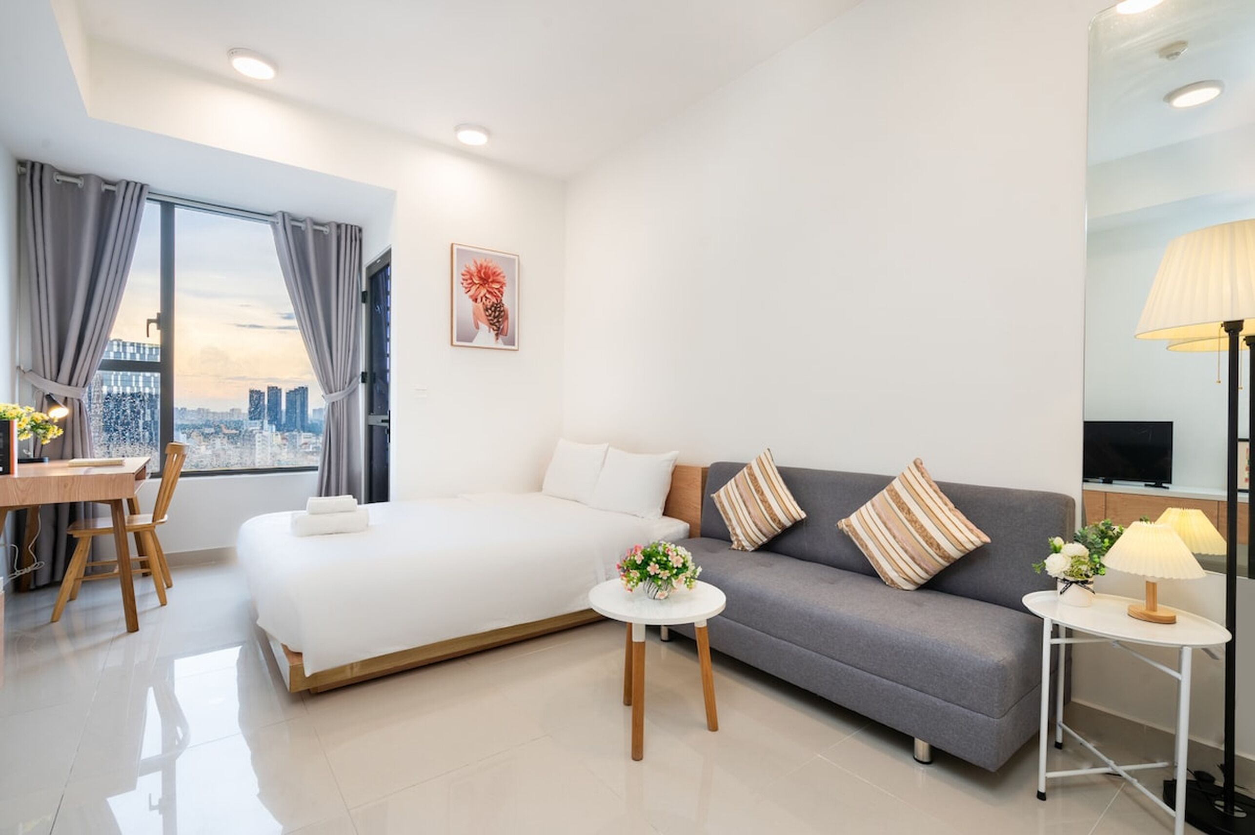 A modern hotel room with a large bed, a gray sofa, a small round table, and a desk with a chair.