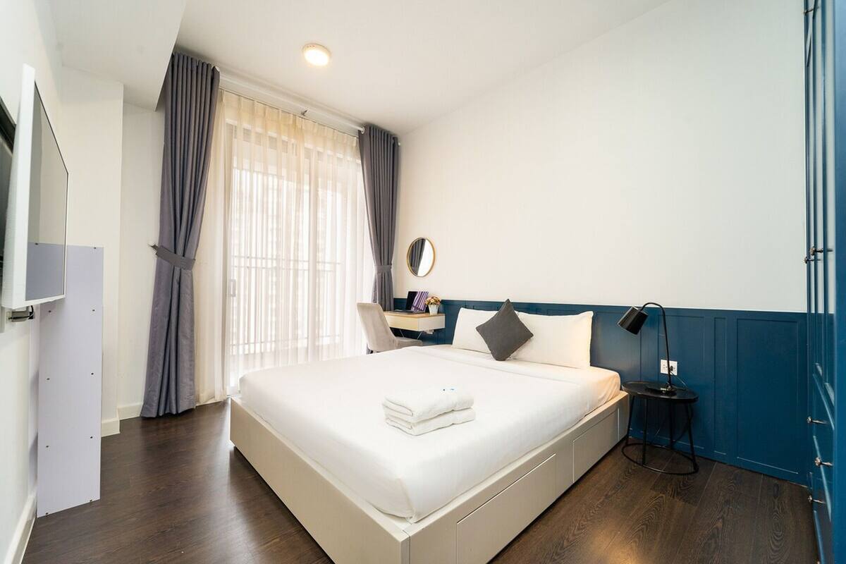 Habitación de hotel ordenada con una cama grande, un escritorio con una silla, un espejo y una mesita de noche con una lámpara.