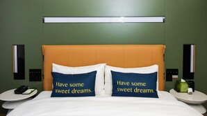 Quarto de hotel com uma cama, duas almofadas com texto, mesa de cabeceira com telefone e luminária na parede.