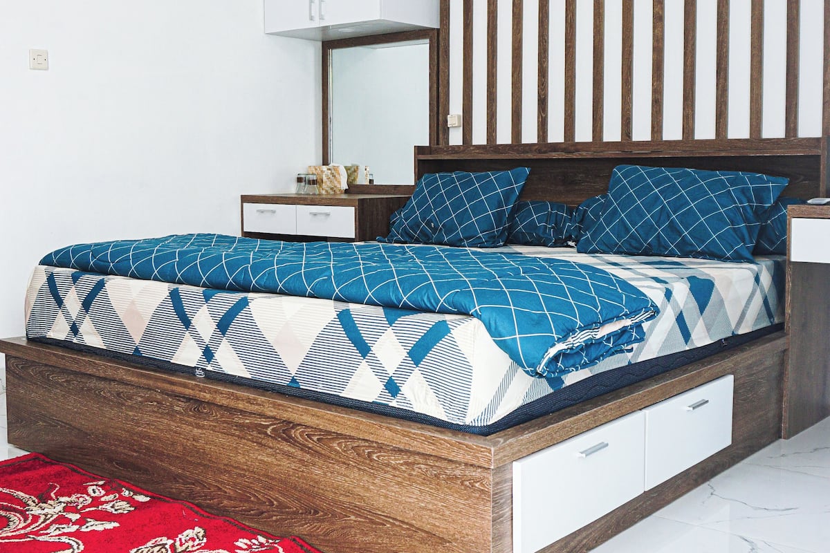 A modern bedroom with a wooden headboard, a patterned bedspread, a red and gold area rug, and a white nightstand with a lamp.