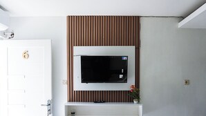 A room with a mounted TV on a wooden panel wall, a door with a number, and a small shelf with a potted plant. - GRIYA GRACHIKA by DOORKOST (Sewon)
