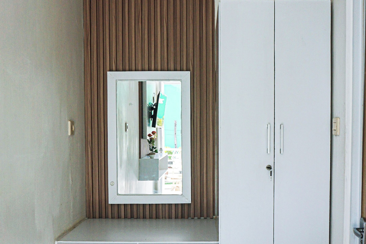 A room with a white desk, a mirror, a wardrobe, a trash bin, and a door.