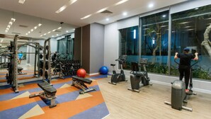 Un gimnasio moderno con diversos equipos de ejercicio, un espejo grande y vistas a un paisaje tropical a través de las ventanas.