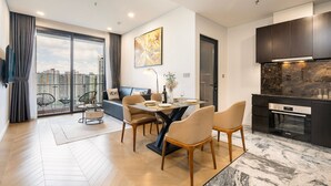 A modern living room with a dining area, a kitchen with a stone backsplash, and a large window with a city view. - Lumiere Riverside - Luxury Condo District 2 (Ho Chi Minh City)