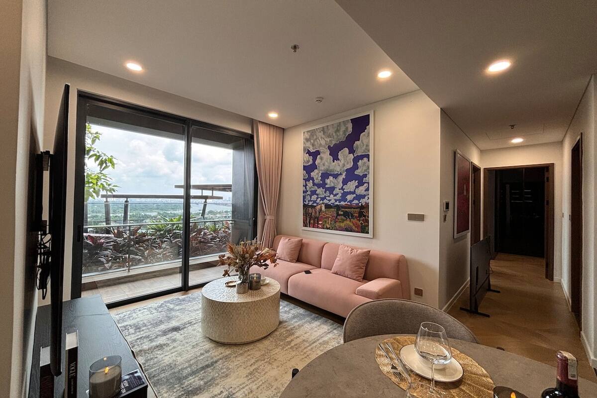 A modern living room with a sofa, a coffee table, and a dining table set for two. A large window offers a view of the outdoors.