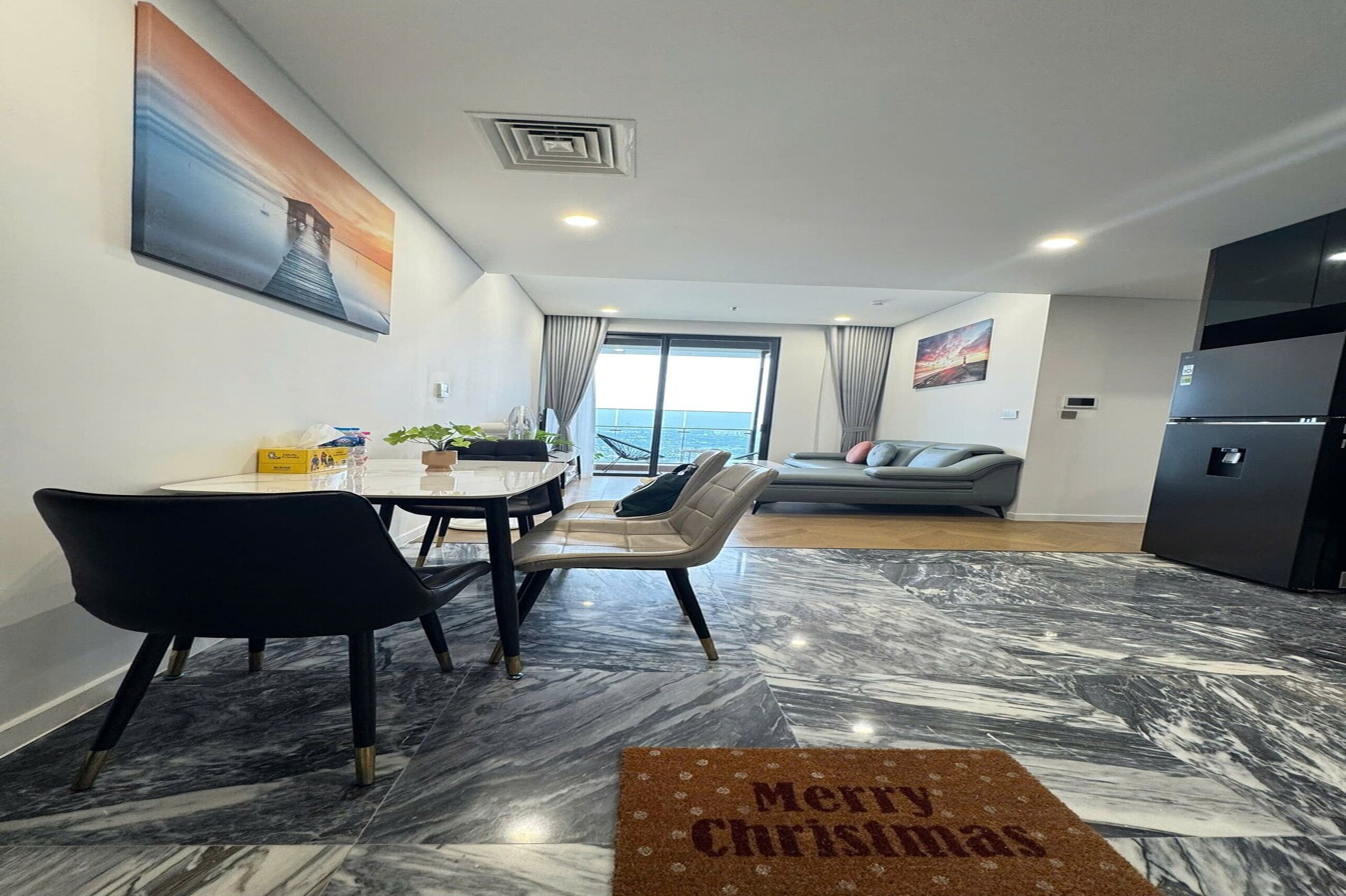 A modern living room with a dining area, a sofa, a large window with a view, and a kitchen area.
