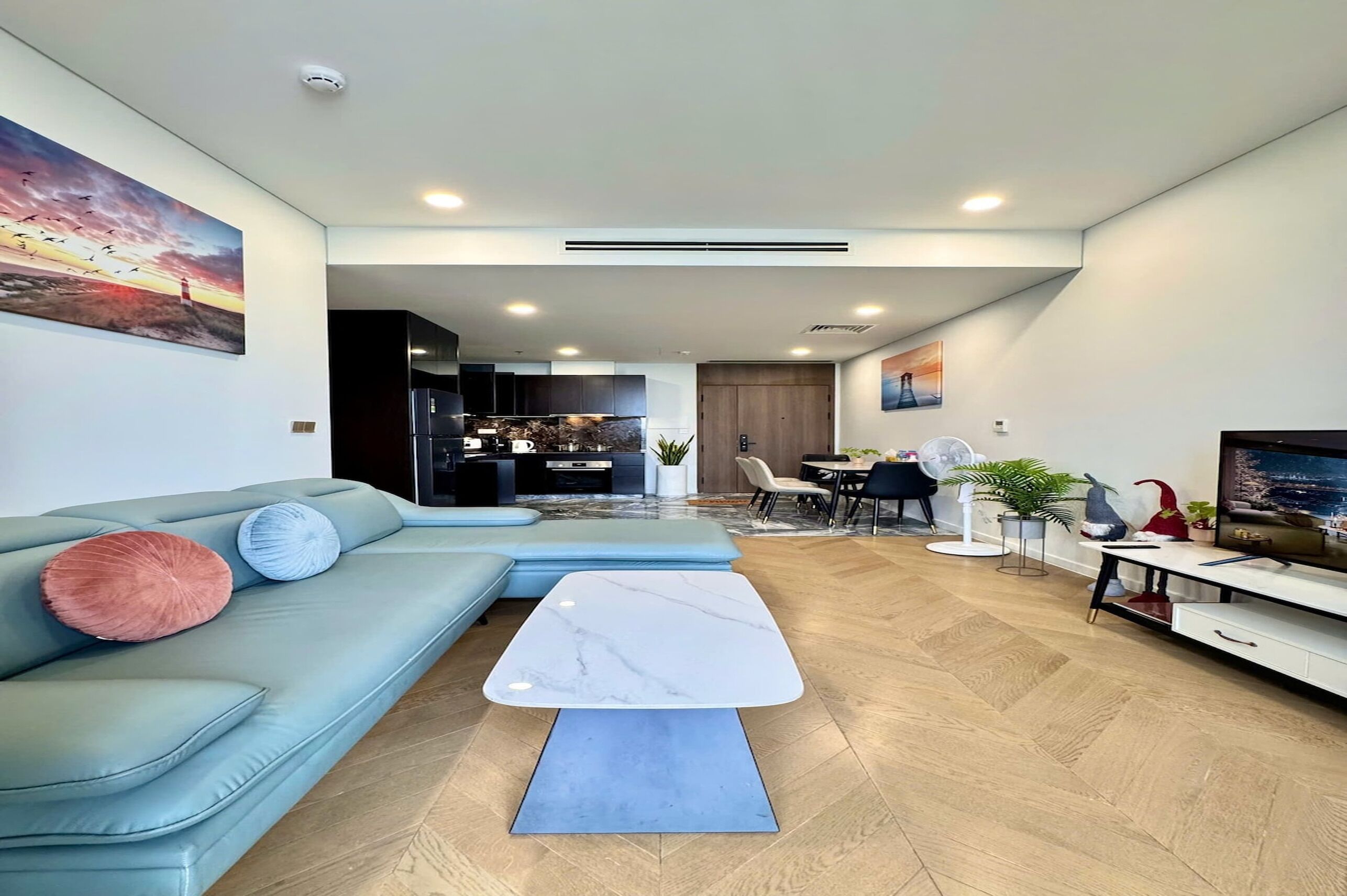 A modern living room with a sectional sofa, a marble coffee table, and a dining area with a TV and plants.