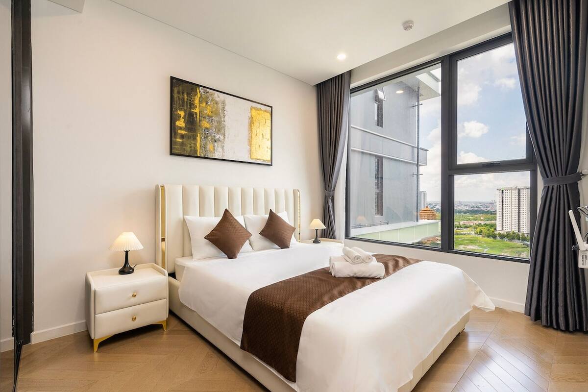 A hotel room with a large bed, a nightstand, a lamp, a painting on the wall, and a view of the city through a large window.