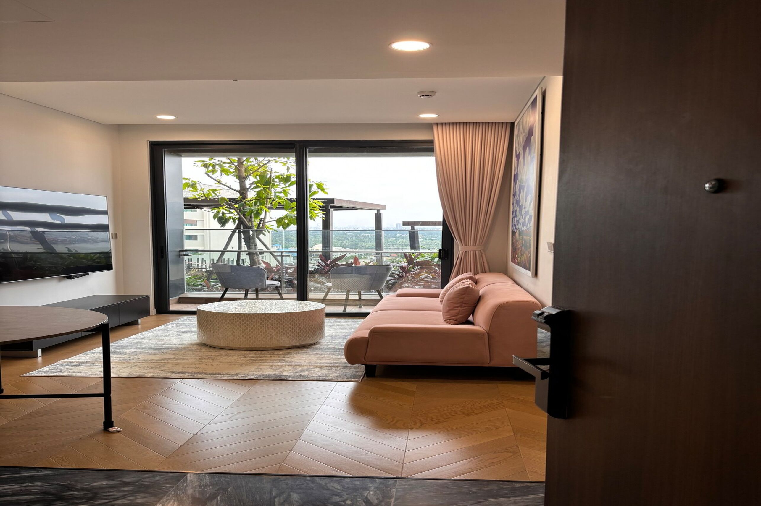 A modern living room with a sofa, a coffee table, and a TV. There is a balcony with a view of buildings and trees.