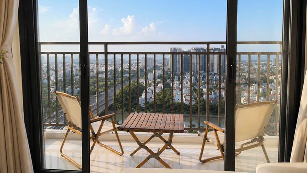 A balcony with a wooden table and two chairs, overlooking a cityscape with buildings and greenery. - The Sun Avenue - Lalaland & Chill Sky Pool (Ho Chi Minh City)