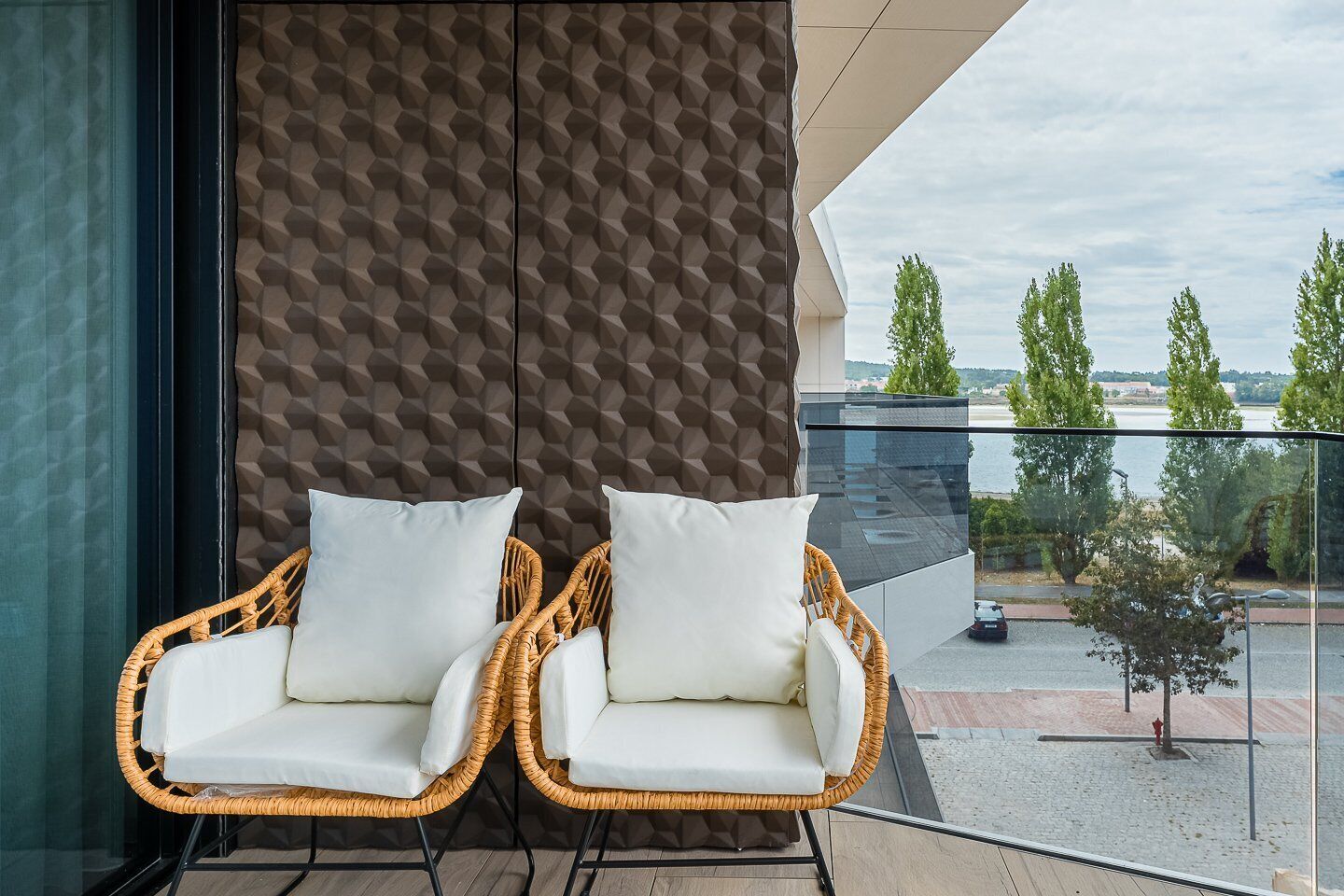 Two wicker chairs with white cushions on a balcony with a geometric wall and a view of trees and water.