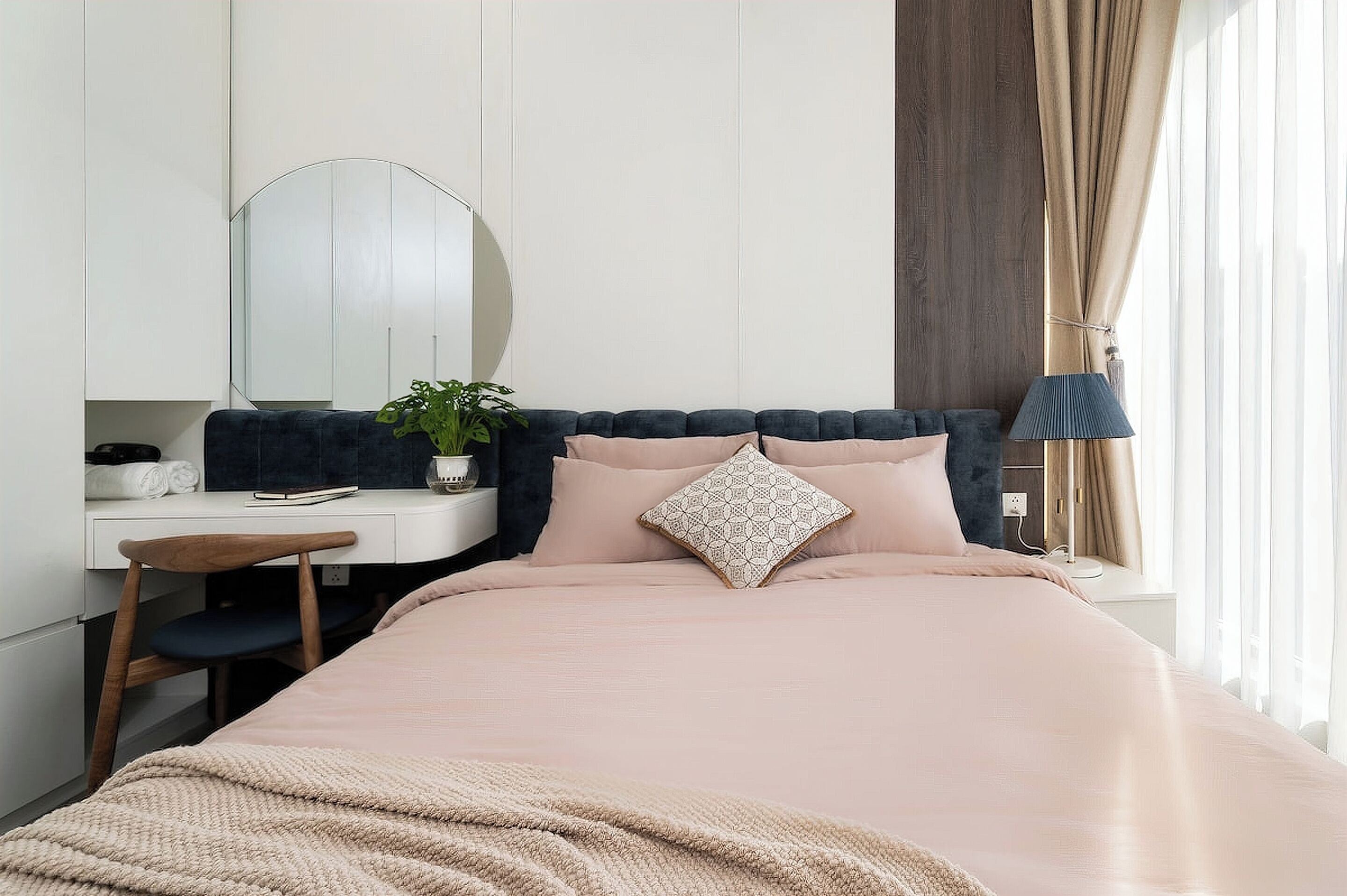 A bedroom with a bed, a desk, a chair, a mirror, a plant, and a window with curtains.