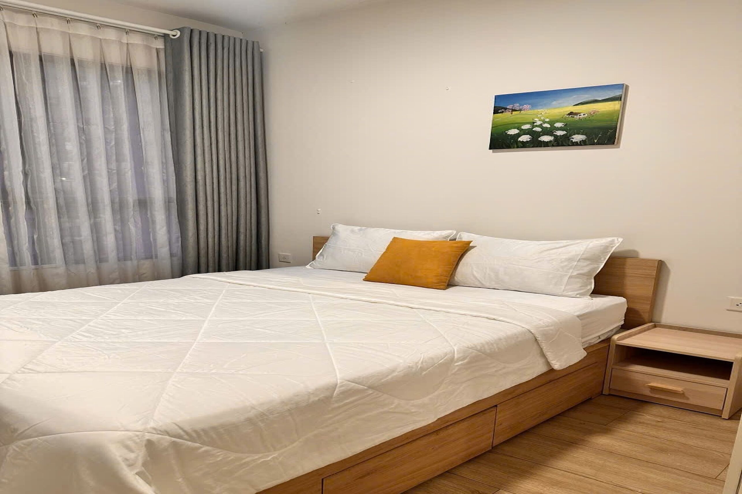 A bedroom with a bed, a nightstand, a painting, and a window with curtains.