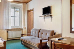 A room with a sofa, a table, a television mounted on the wall, and a window with curtains.