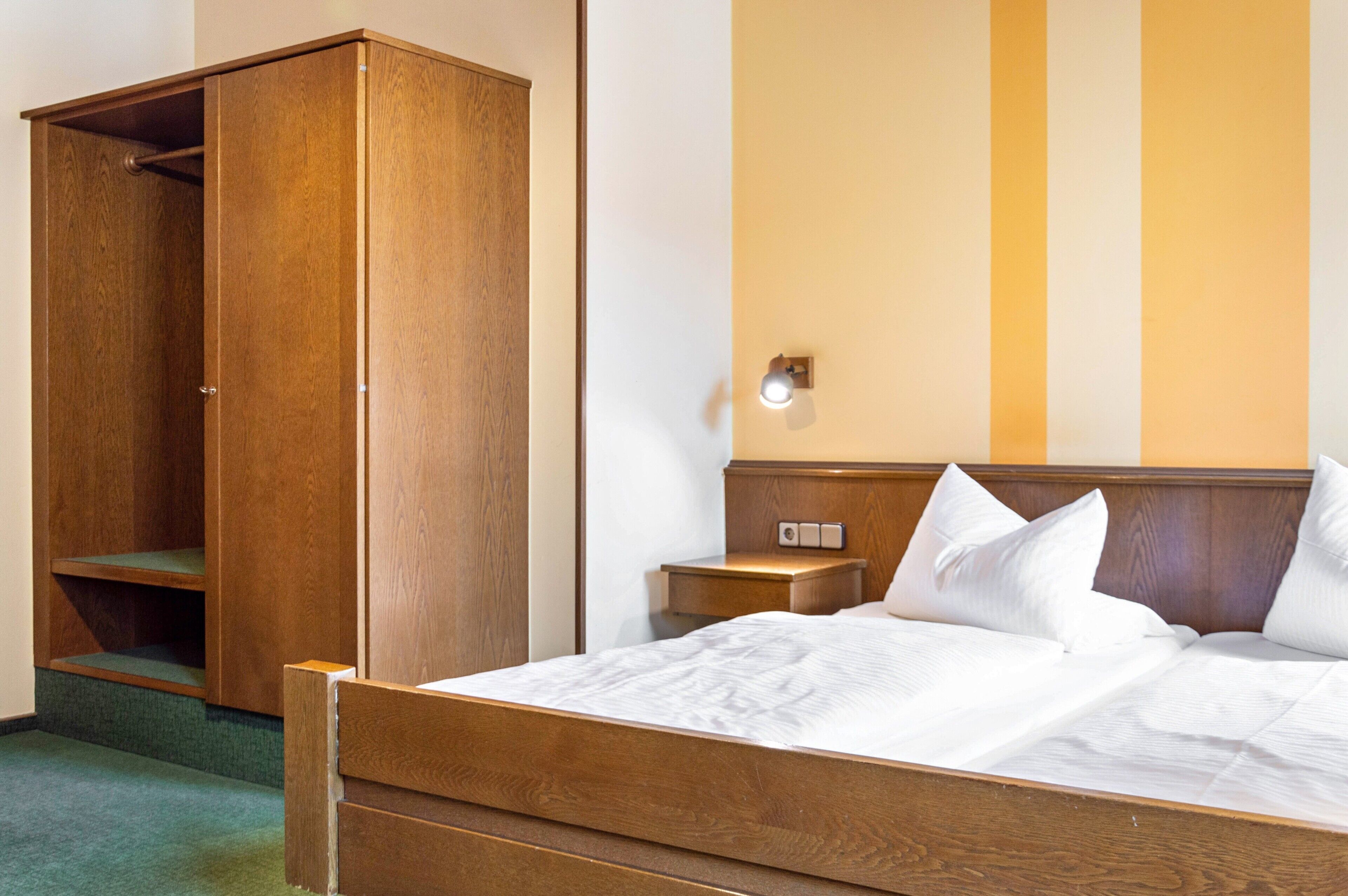 Une chambre d’hôtel avec un lit en bois, une armoire, une table de chevet et une lampe fixée au mur.