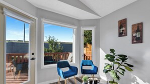 A bright room with two blue chairs, a small table with a plant and mugs, and a view of a wooden deck and trees through large windows.