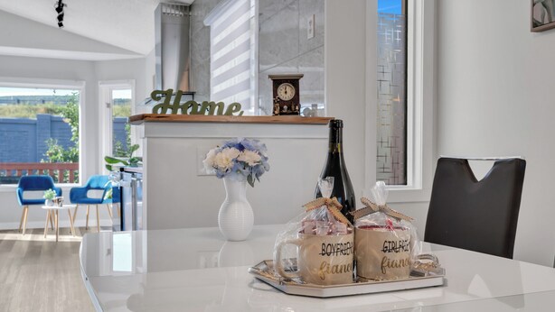 A modern kitchen with a dining area. There is a tray with two mugs labeled "boyfriend" and "girlfriend" and a bottle of champagne. A clock and a vase with flowers are also visible.
