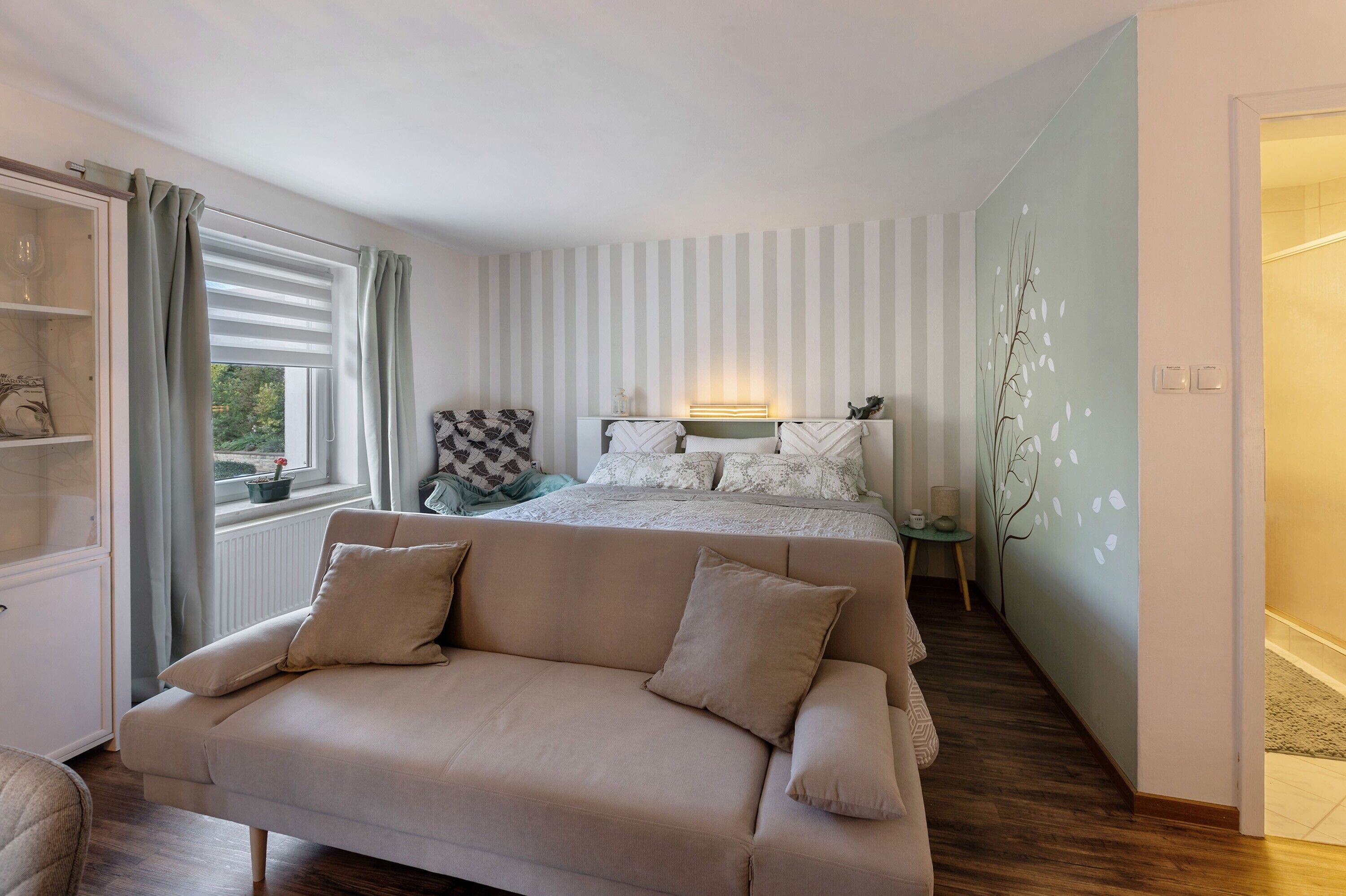 Une chambre à coucher comprenant un lit, un canapé, une fenêtre avec des rideaux, une table de chevet et un mur décoré d’un motif représentant un arbre.