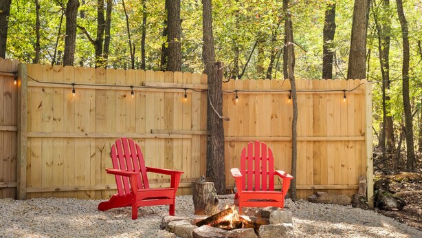 Two red Adirondack chairs facing a fire pit surrounded by pebbles, with a wooden fence and string lights in the background. - Luxe 2 Bed Dome w Hot Tub, Deck, Fire Pit & Bath (Augusta)