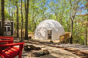 A dome-shaped cabin in a forest with a wooden deck and a red chair.