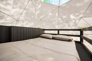 Una cama con colcha blanca y dos almohadas, cabecera de madera y vistas a través de una estructura de ventana geométrica.