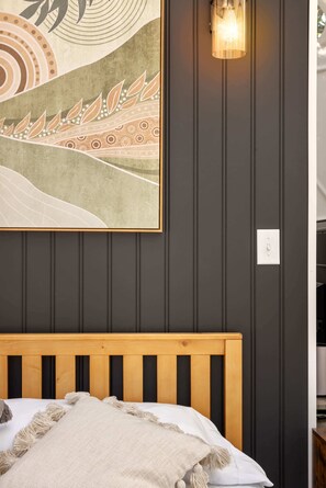 Un dormitorio con cabecera de madera, cama con manta blanca y beige, lámpara de pared y un cuadro abstracto grande.