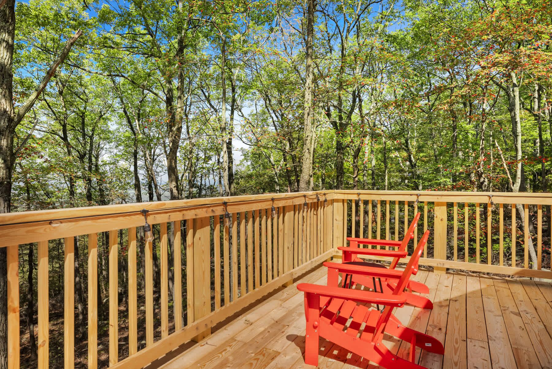 A wooden deck with a railing, two red Adirondack chairs, and a forested backdrop.
