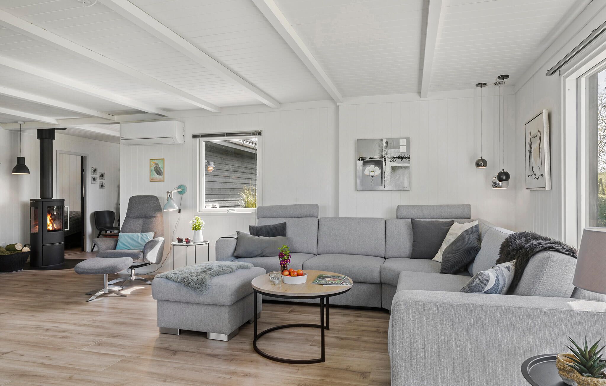 Un salon moderne avec un canapé gris, un pouf et une table basse. On aperçoit une cheminée dans un coin, et une fenêtre laisse entrer la lumière naturelle.