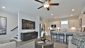 A modern living room with a TV, sofa, and coffee table. There's a kitchen with a bar and island, and a staircase leading to another level.