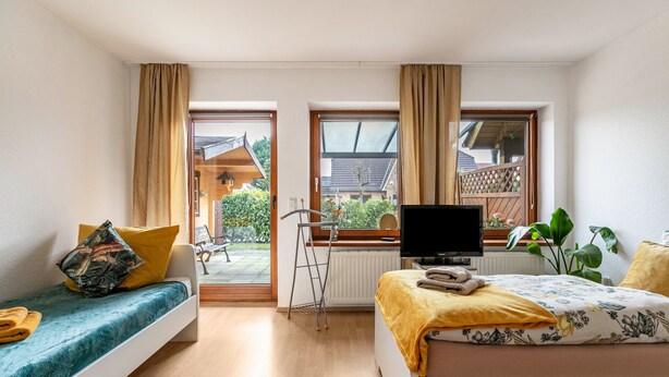 A bedroom with a bed, a desk with a computer, a window with curtains, and a view of the outside.