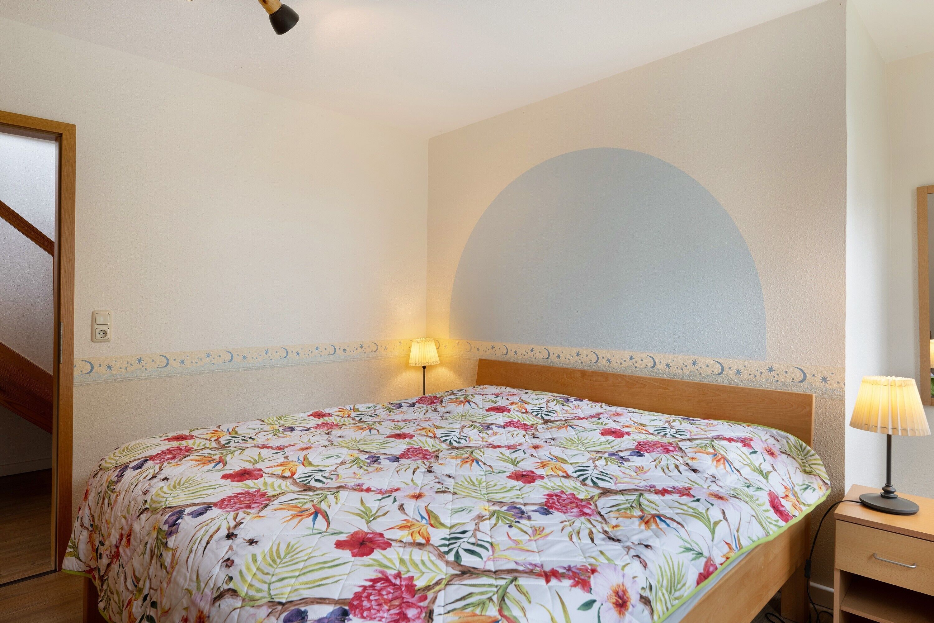 A bedroom with a floral bedspread, a nightstand with a lamp, a wooden headboard, and a large arched wall decoration.