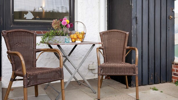 Twee rieten stoelen, een tafel en een mand met fruit en bloemen voor een gebouw met een zwarte deur en een raam met een vissen silhouet.