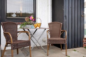 Twee rieten stoelen, een tafel en een mand met fruit en bloemen voor een gebouw met een zwarte deur en een raam met een vissen silhouet.