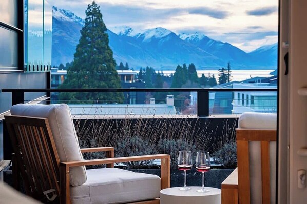 A balcony with a view of mountains and a lake, a chair, a small table with glasses, and a railing.