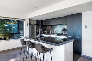 A modern kitchen with a large island, bar stools, dark cabinetry, and a view of greenery outside.