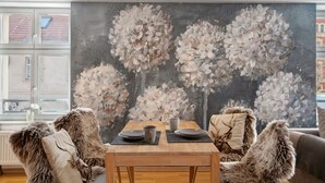 Ein Essbereich mit einem für zwei Personen gedecktem Tisch, einem großen Blumenbild an der Wand und Blick auf ein Gebäude durch ein Fenster.
